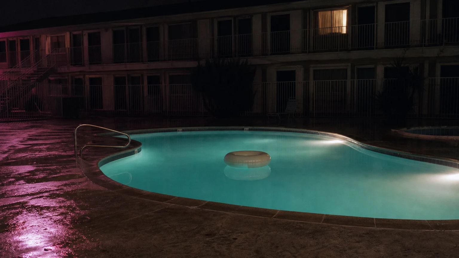 An empty motel pool lit turquoise at night, one inflatable ring drifting, a neon vacancy sign reflecting pink on the wet deck.