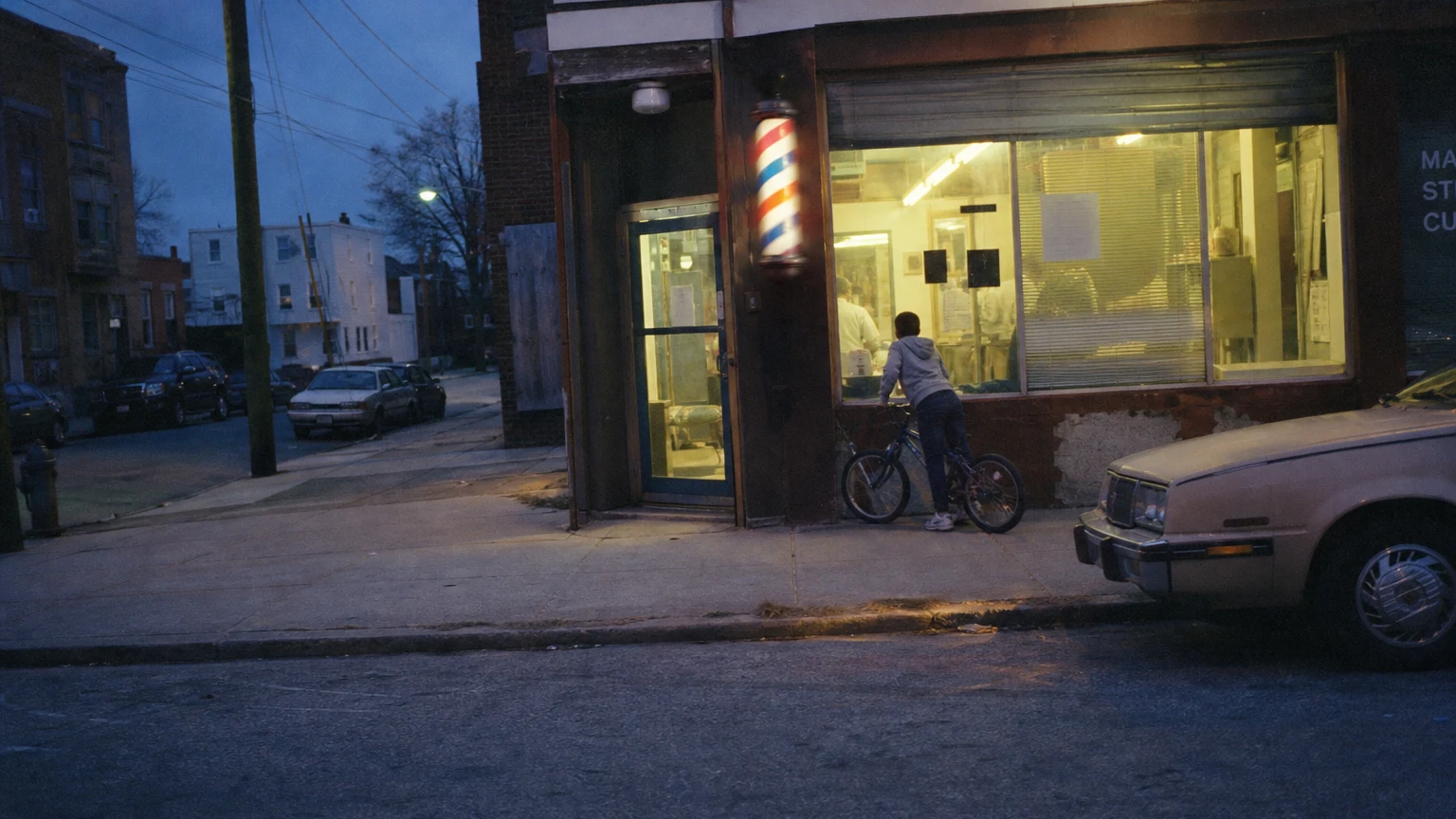 A neighborhood barbershop at dusk from across the street, barber pole lit, a kid on a bike looking in.