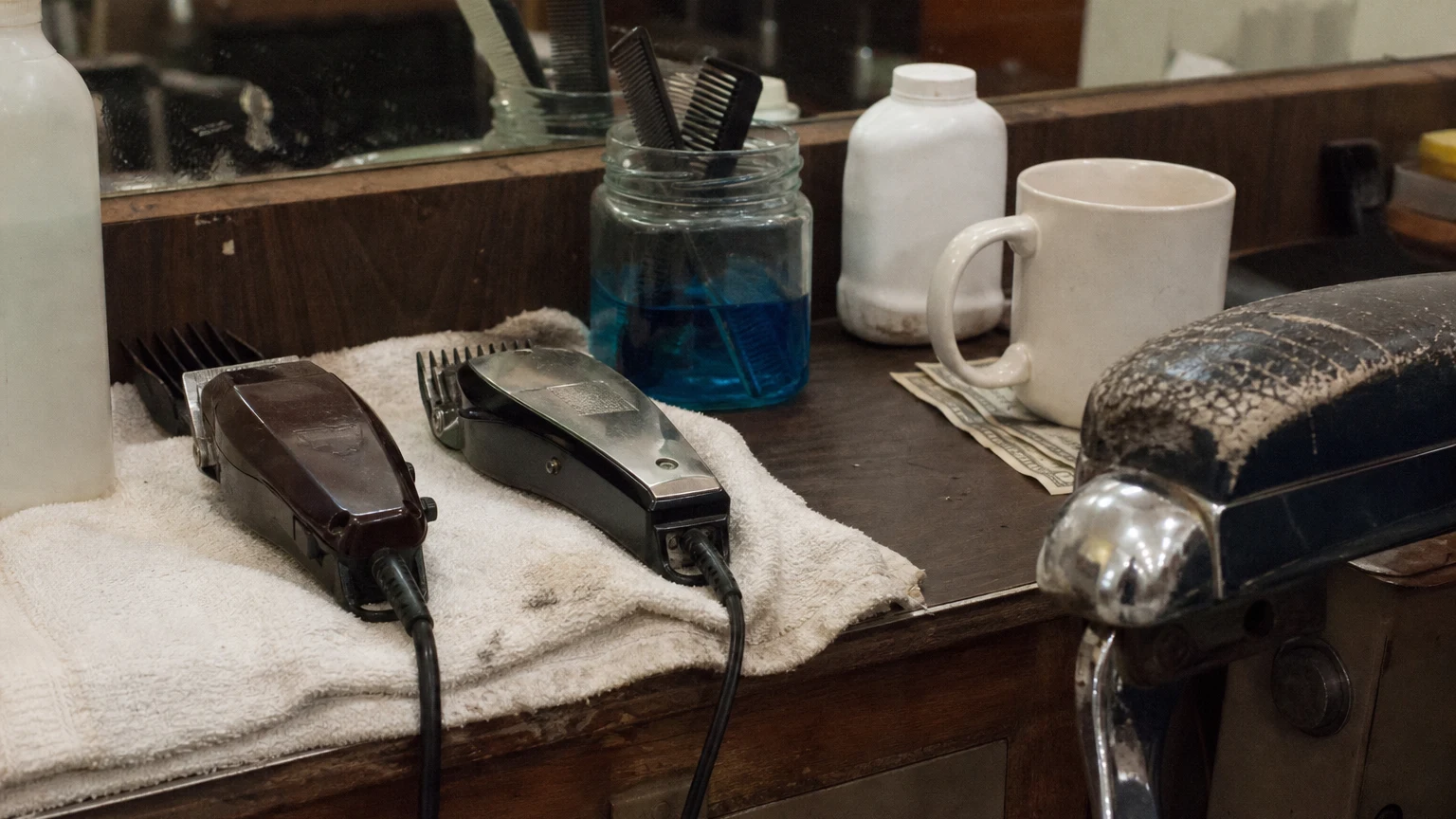 A counter detail in the same shop: clippers on a folded towel, combs in blue disinfectant, bills under a mug.