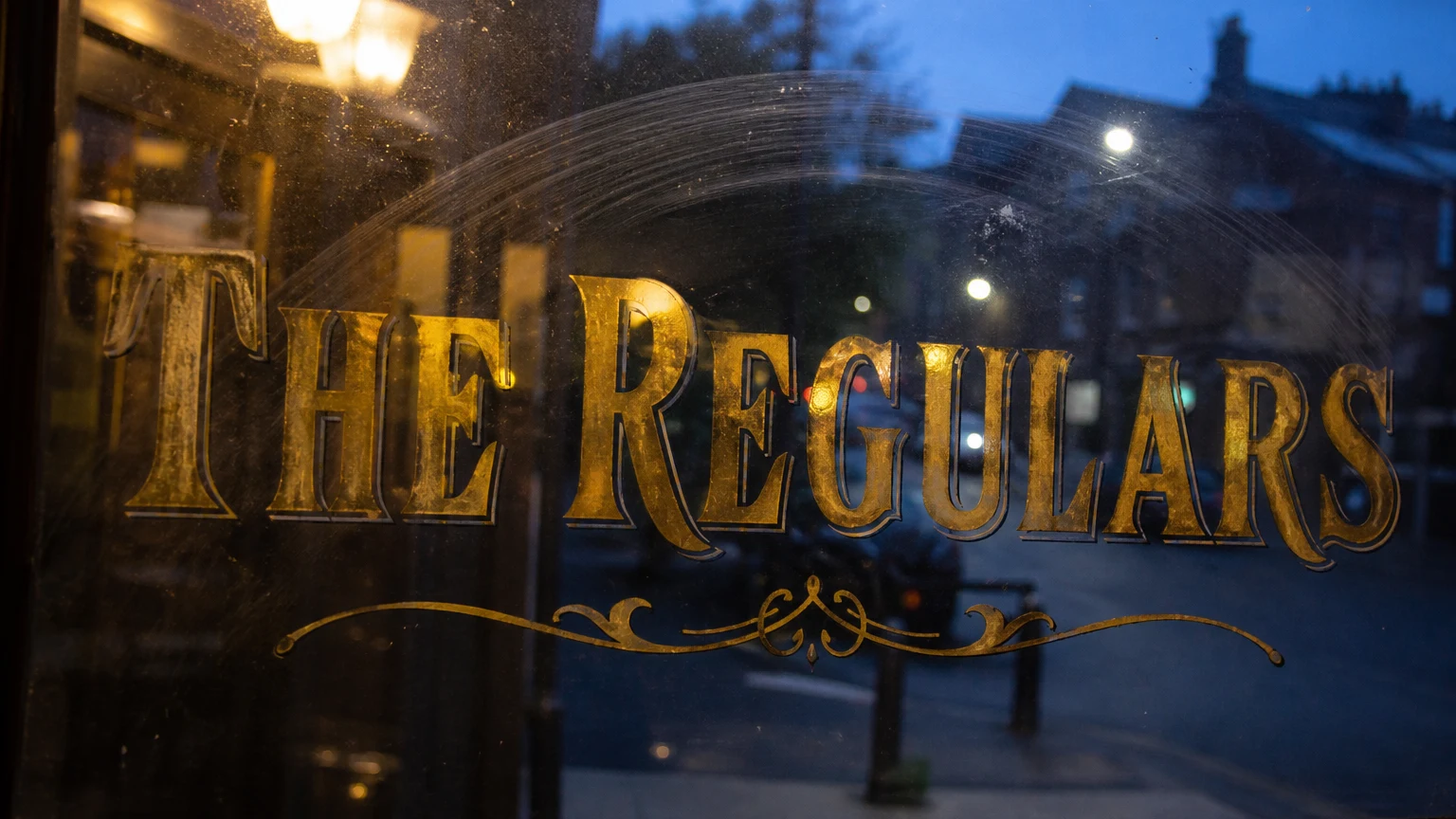 Gold-leaf lettering etched on a pub window at dusk, the street soft and blue beyond the glass.