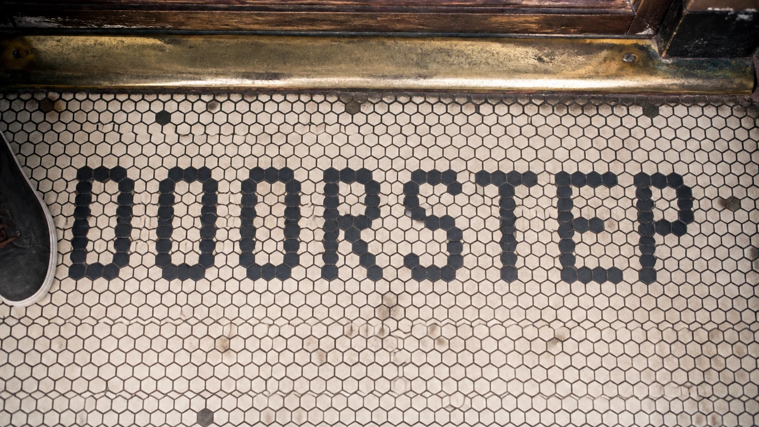 A shop threshold mosaic spelling a title in dark hexagonal tiles on cream, a brass door rail crossing above.